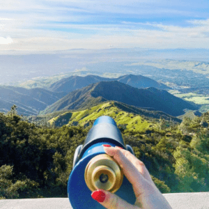 A woman's hand hold a scope that points towards green valleys and hills from Mt. Diablo State Park